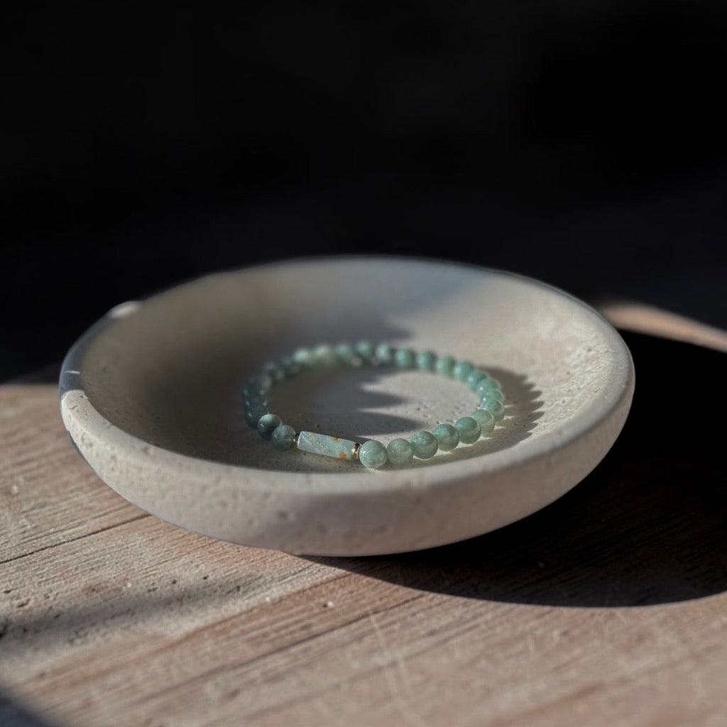 A beaded bracelet in pale mint green rests inside a small round ceramic dish on a wooden surface, with dramatic sunlight and shadows.