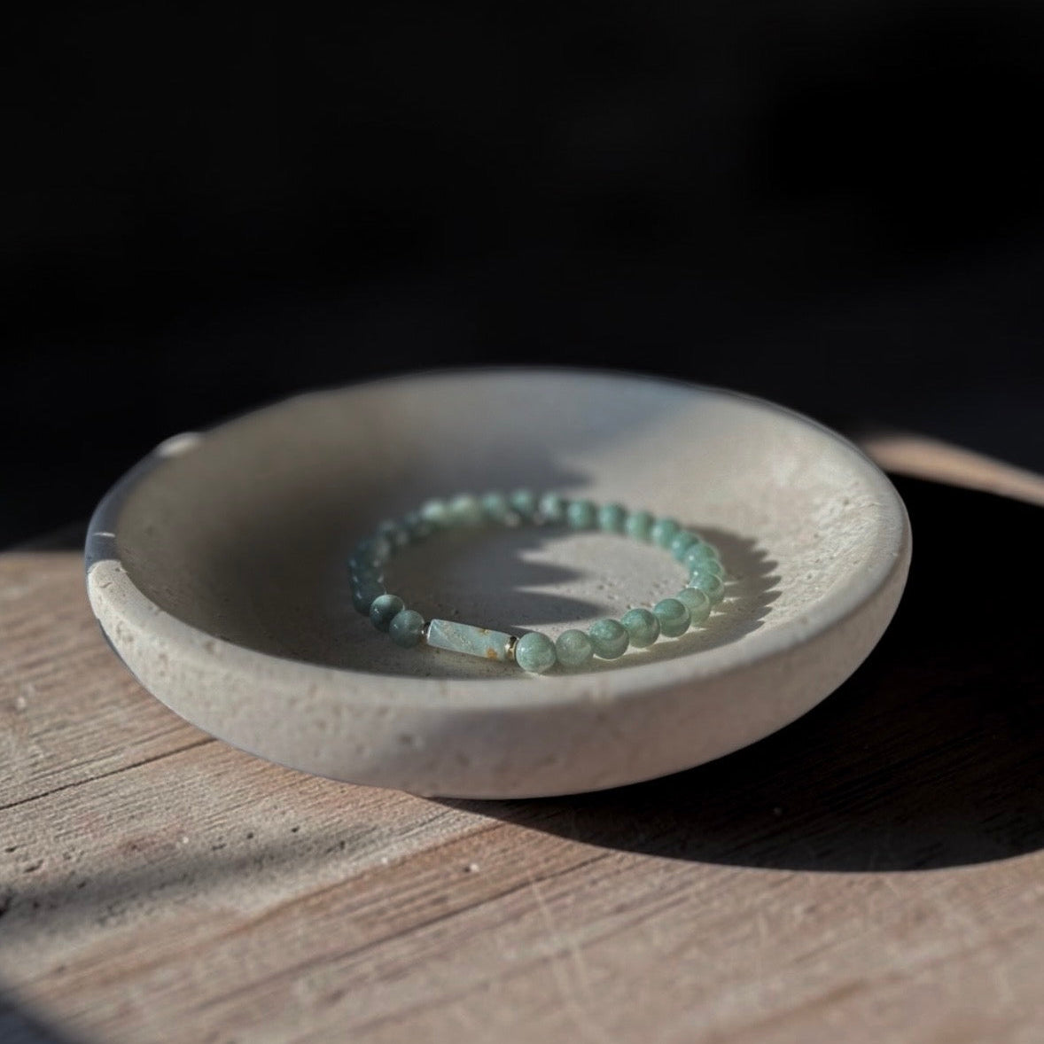 A beaded bracelet in pale mint green rests inside a small round ceramic dish on a wooden surface, with dramatic sunlight and shadows.