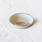 A beaded bracelet with peach-pink round beads and a small mint green rectangular accent, displayed in a shallow round ceramic dish on a textured off-white surface.