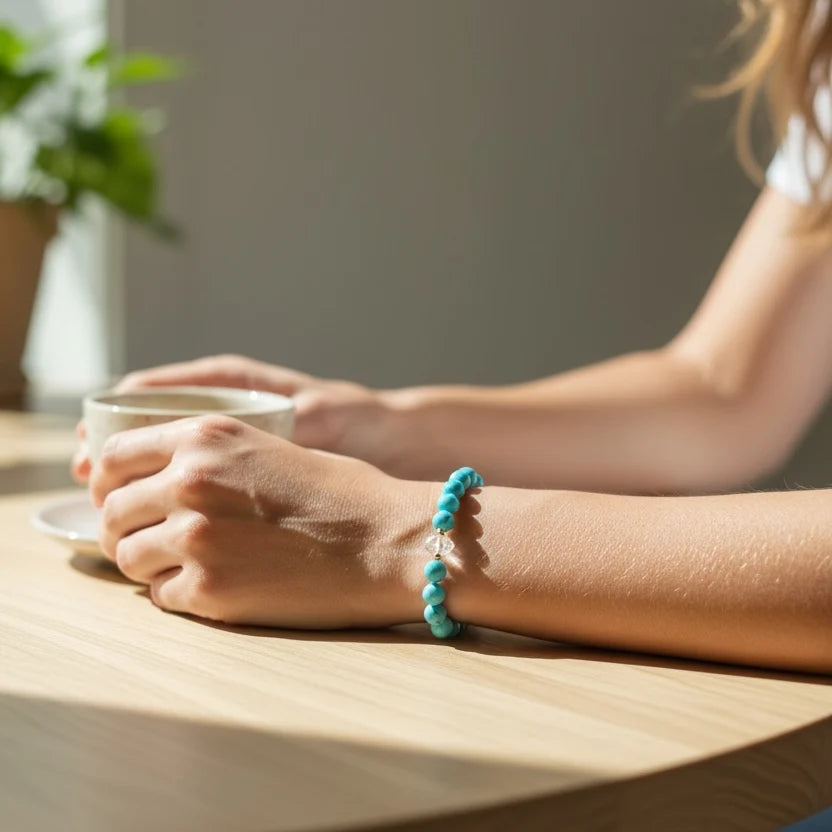 A turquoise beaded bracelet with a small gold clasp worn on a person's wrist, shown in a warm, softly lit indoor setting with a wooden table and a cup in the background.