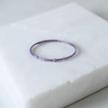 A delicate beaded bracelet in shades of purple and lavender, featuring small metallic silver beads, displayed on a white marble block.