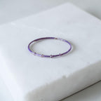 A delicate beaded bracelet in shades of purple and lavender, featuring small metallic silver beads, displayed on a white marble block.