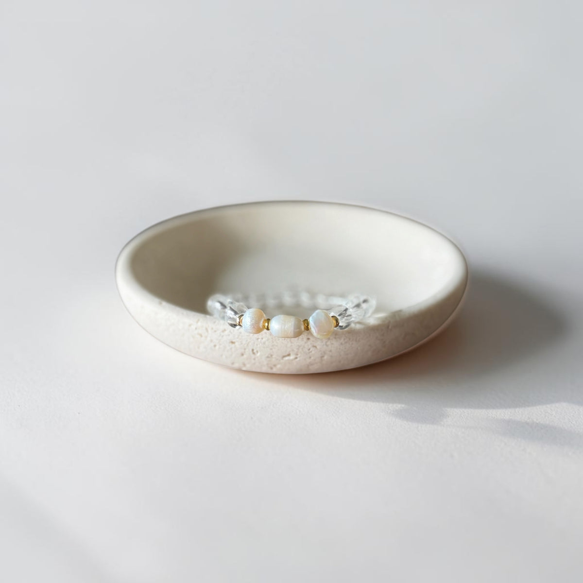 A clear beaded bracelet with small pearl-like beads rests inside a shallow, textured off-white ceramic dish on a clean white surface.