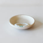 A beaded stretch bracelet with white round beads and a small light blue rectangular accent, displayed inside a small speckled cream ceramic dish on a light gray background.
