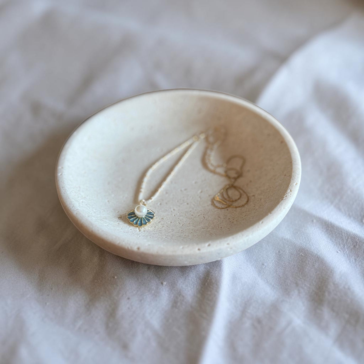 A small round ceramic jewelry tray in a warm off-white speckled finish, holding a delicate necklace with a small blue-green and gold pendant, set on a soft white fabric background.