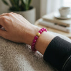 A close-up of a person's wrist wearing a beaded bracelet made of magenta pink round beads with a single polished gold accent bead in the center, styled with a black ribbed sleeve against a cozy indoor background.