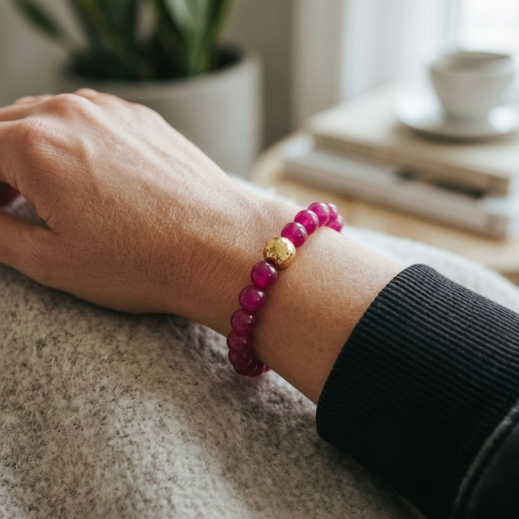 A close-up of a person's wrist wearing a beaded bracelet made of magenta pink round beads with a single polished gold accent bead in the center, styled with a black ribbed sleeve against a cozy indoor background.