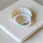 A beaded bracelet with white round beads and a small mint green rectangular accent, displayed on a scalloped cream dish atop a textured beige stone block.