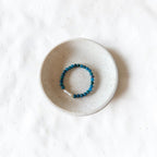 A beaded stretch bracelet in deep teal blue with a small white stone accent, displayed in a small speckled ceramic dish on a textured white background.