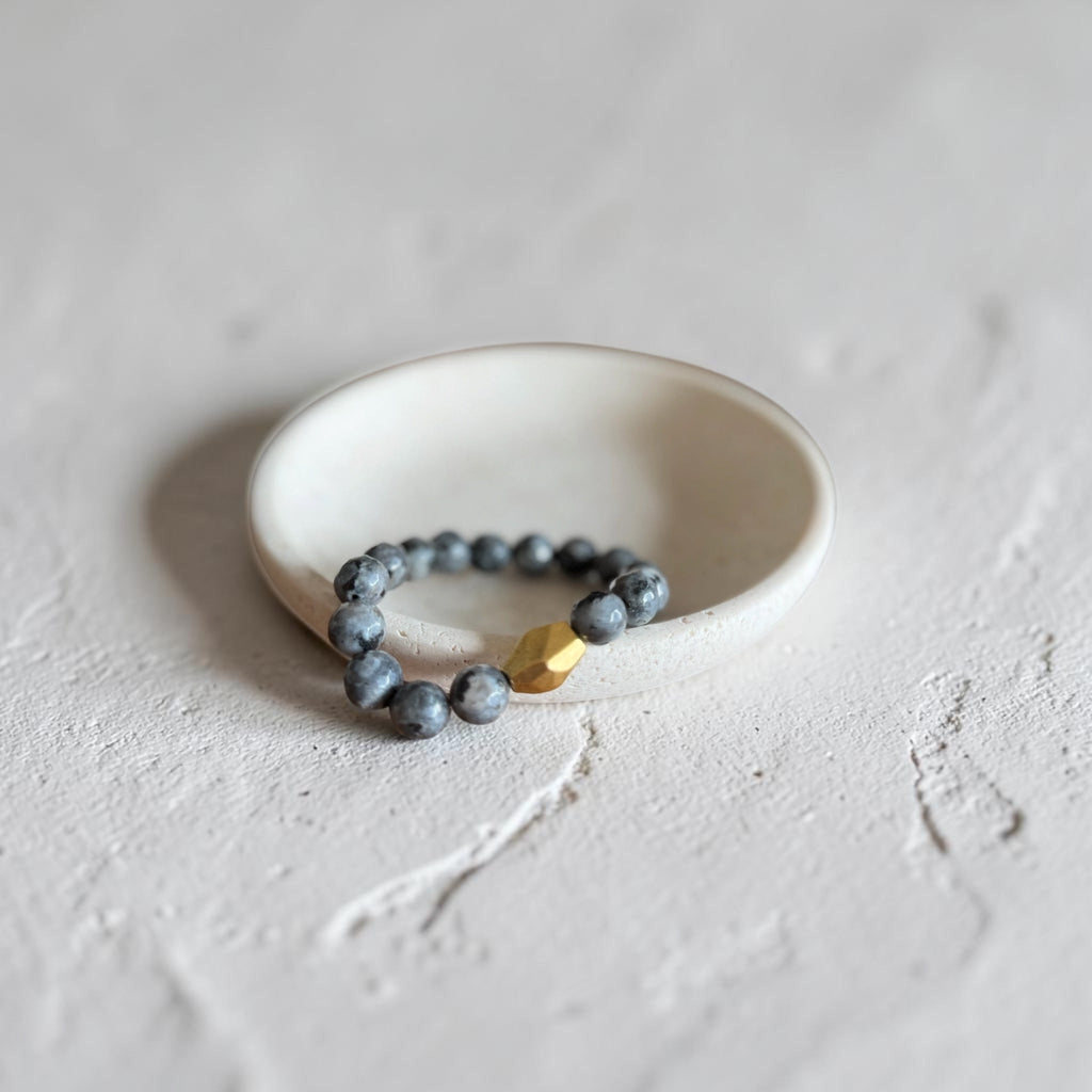 A beaded stretch bracelet with dark gray stone beads and a single faceted gold accent bead, displayed on a small white ceramic dish against a textured light gray background.