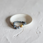 A beaded stretch bracelet with dark gray stone beads and a single faceted gold accent bead, displayed on a small white ceramic dish against a textured light gray background.