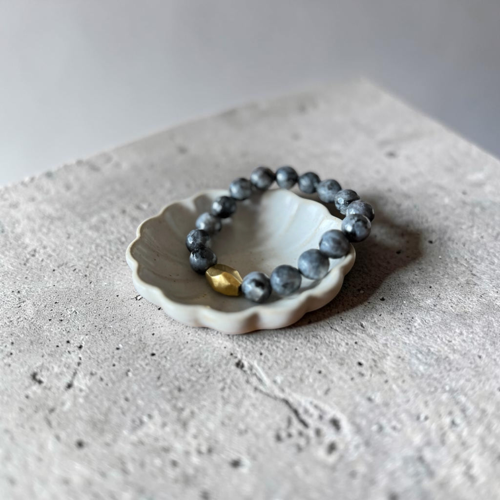 A beaded bracelet with dark gray-blue round beads and a small gold accent bead, displayed on a small white ceramic dish over a textured gray stone surface.