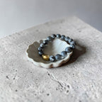 A beaded bracelet with dark gray-blue round beads and a small gold accent bead, displayed on a small white ceramic dish over a textured gray stone surface.