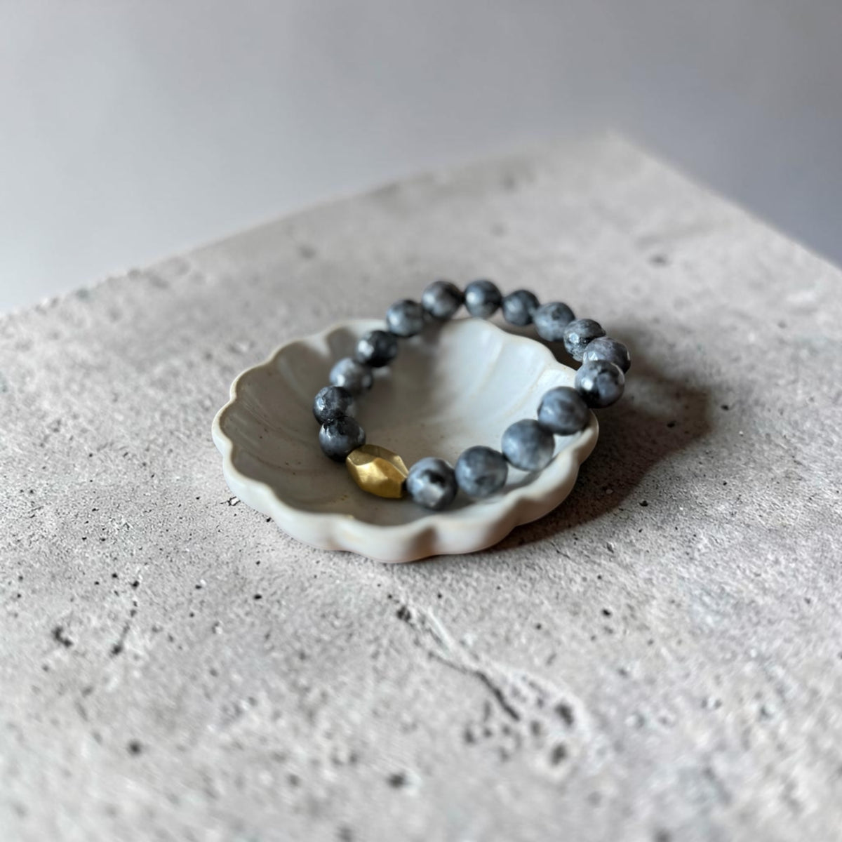 A beaded bracelet with dark gray-blue round beads and a small gold accent bead, displayed on a small white ceramic dish over a textured gray stone surface.
