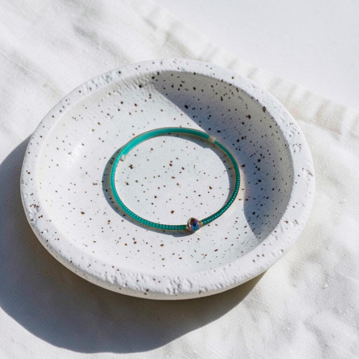 A thin turquoise green beaded bracelet with a small iridescent bead, displayed inside a white speckled ceramic bowl on a white fabric background.