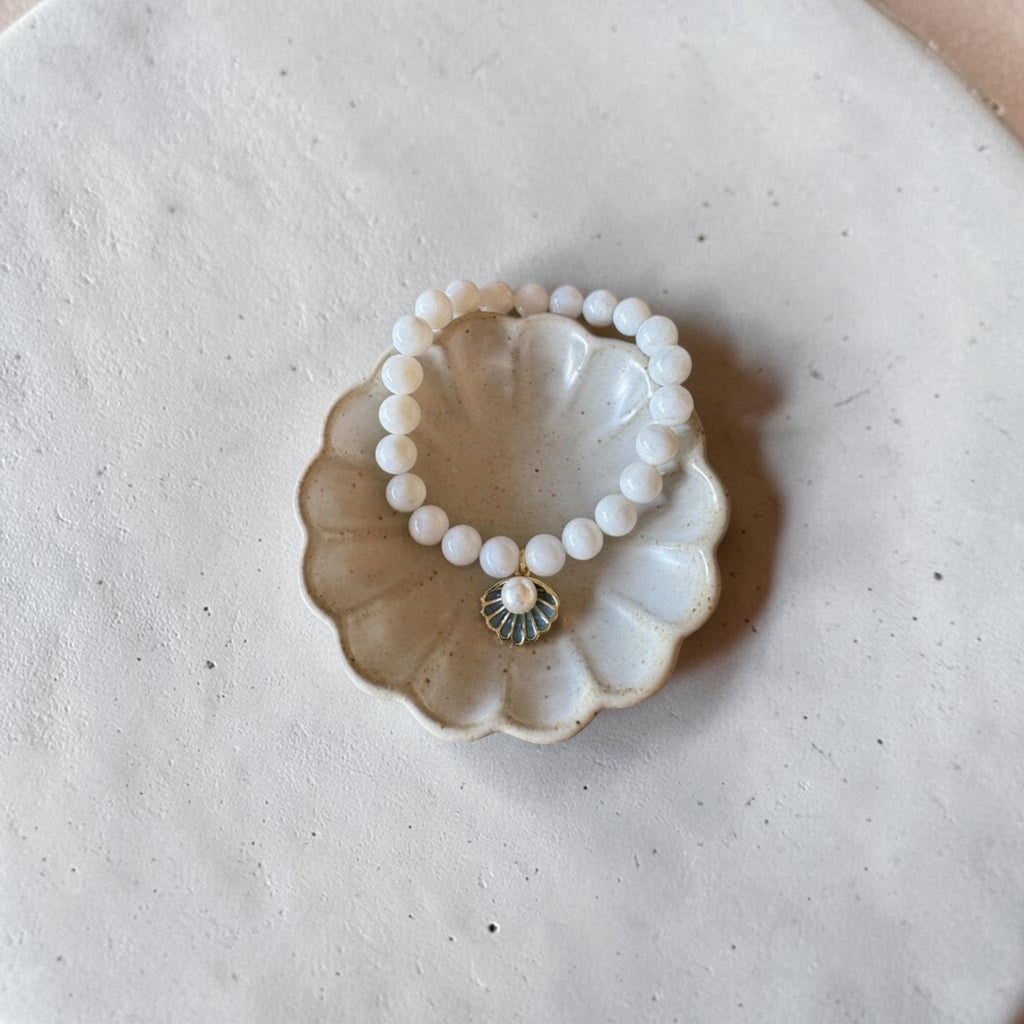 A beaded stretch bracelet with white round beads and a small gold-toned sunburst charm, displayed on a scalloped ceramic dish over a textured off-white surface.