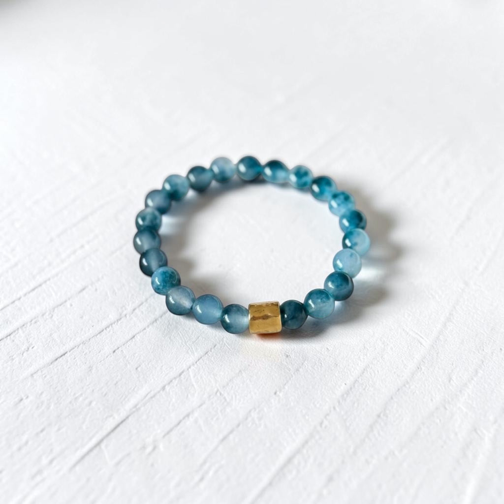 A beaded stretch bracelet made of translucent blue-gray beads with a small gold-toned rectangular accent bead, photographed on a white textured surface.
