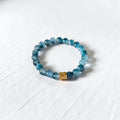A beaded stretch bracelet made of translucent blue-gray beads with a small gold-toned rectangular accent bead, photographed on a white textured surface.