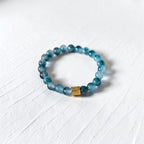 A beaded stretch bracelet made of translucent blue-gray beads with a small gold-toned rectangular accent bead, photographed on a white textured surface.