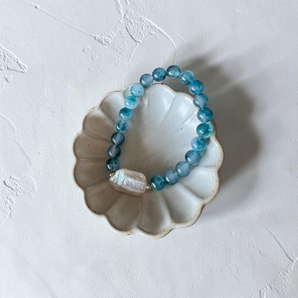 A beaded stretch bracelet with translucent blue-green round beads and a textured white stone accent, displayed on a small white scalloped dish against a textured off-white background.