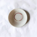 A small round ceramic saucer with a speckled off-white glaze and a thin red circular line inside, photographed from above on a white textured surface.