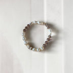 A beaded stretch bracelet featuring white baroque pearls, translucent brown smoky beads, and small metallic gold spacer beads, displayed on a light beige textured background.