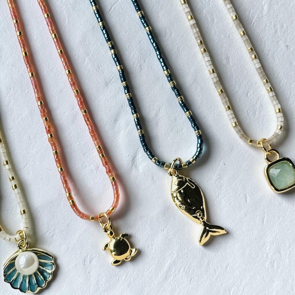 Four delicate beaded necklaces with gold-tone pendants are arranged diagonally on a textured white surface. The necklaces feature coral pink, metallic blue, and cream beads, with pendants including a gold fish, a gold turtle, a blue and white seashell with a pearl, and a light green stone.