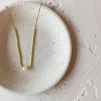 A delicate gold chain necklace with a single white pearl pendant, displayed on a round, textured beige ceramic dish against a matching beige background.
