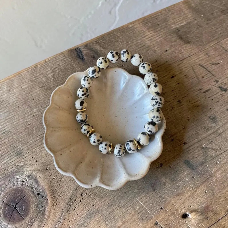 A beaded stretch bracelet with white beads speckled in black, displayed on a small scalloped ceramic dish over a rustic wooden surface.