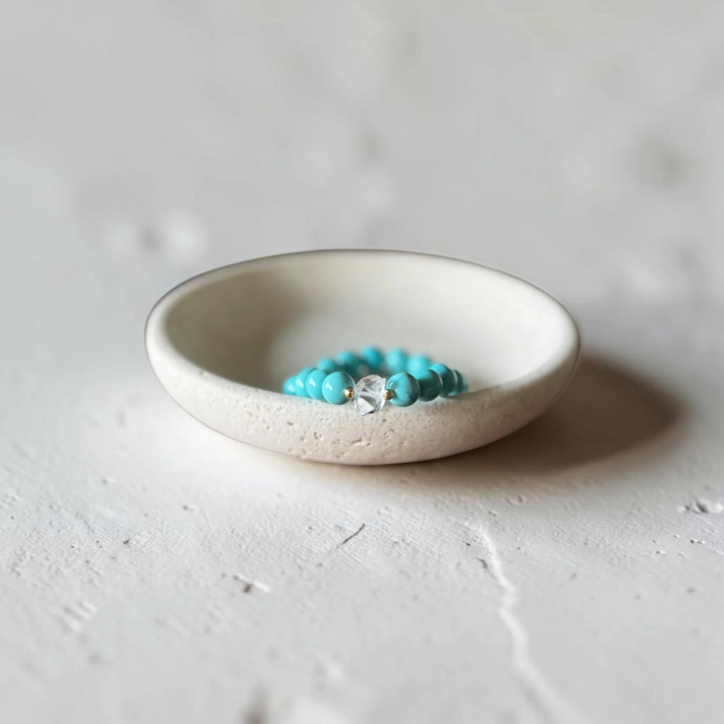 A small turquoise beaded bracelet with a clear faceted center stone, resting inside a shallow, textured off-white ceramic dish on a light gray surface.