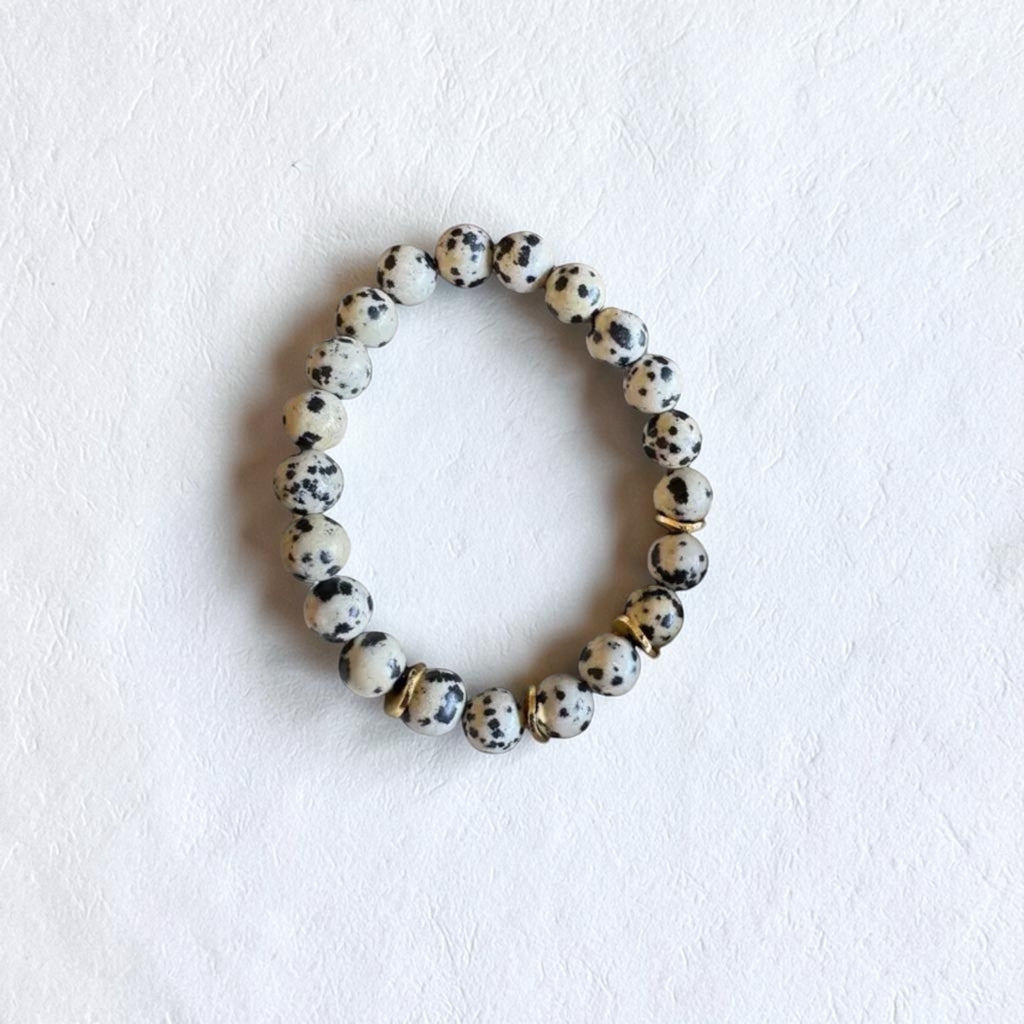 A beaded stretch bracelet made of round white beads with black speckles, accented by a few metallic gold spacer beads, displayed on a textured white surface.