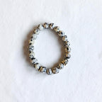 A beaded stretch bracelet made of round white beads with black speckles, accented by a few metallic gold spacer beads, displayed on a textured white surface.