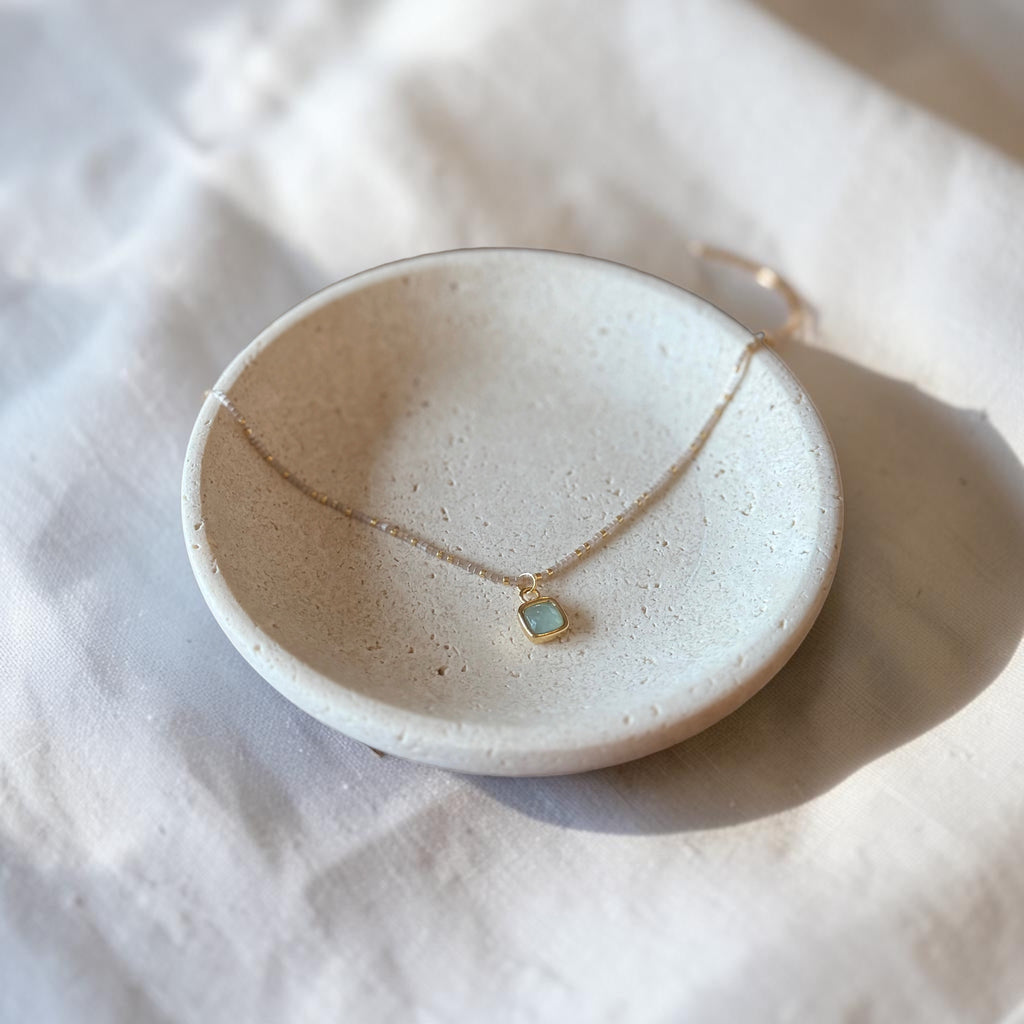 A delicate gold chain necklace with a small square pendant featuring a light blue-green stone, displayed on a small beige ceramic dish over a textured white fabric background.