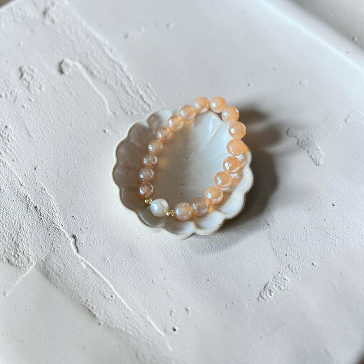 A beaded stretch bracelet with peach-orange translucent beads and a few white pearl-like beads, displayed on a small white scalloped dish over a textured off-white surface.