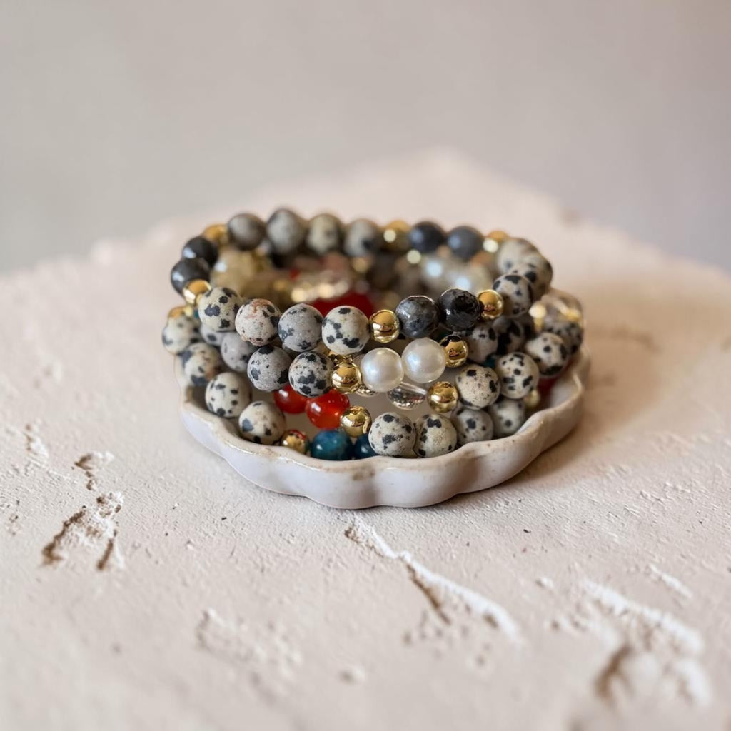 A stack of beaded stretch bracelets displayed on a small white ceramic dish. The bracelets feature white and black speckled dalmatian-style beads, dark gray beads, white pearl-like beads, and small metallic gold and red accent beads.