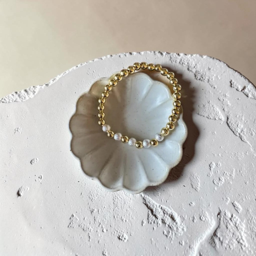 A delicate beaded stretch bracelet with alternating gold and white pearl-like beads, displayed on a small white ceramic shell-shaped dish over a textured beige surface.