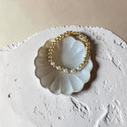 A delicate beaded stretch bracelet with alternating gold and white pearl-like beads, displayed on a small white ceramic shell-shaped dish over a textured beige surface.