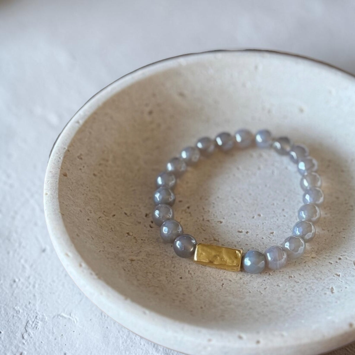 A beaded stretch bracelet with pale gray-blue translucent beads and a small gold rectangular accent, displayed inside a light beige ceramic dish.