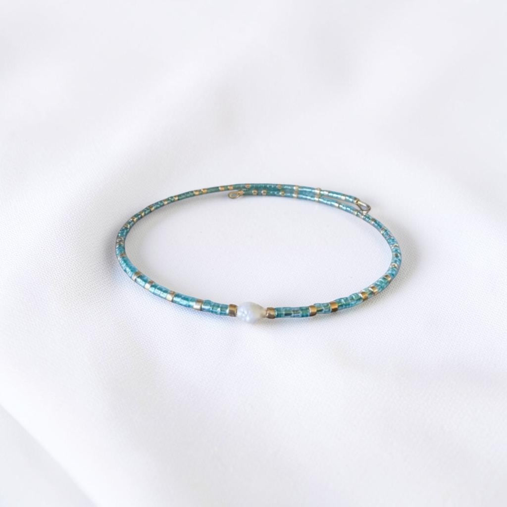 A delicate beaded bracelet with turquoise blue beads, small gold spacer beads, and a single white pearl accent, displayed on a soft white fabric background.
