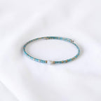 A delicate beaded bracelet with turquoise blue beads, small gold spacer beads, and a single white pearl accent, displayed on a soft white fabric background.