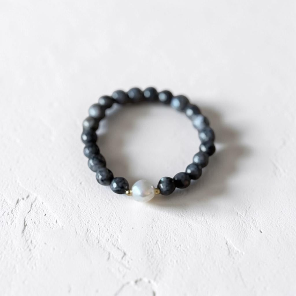 A beaded stretch ring featuring dark gray to black round beads with a single white pearl accent bead and a tiny gold spacer, resting on a textured off-white surface.