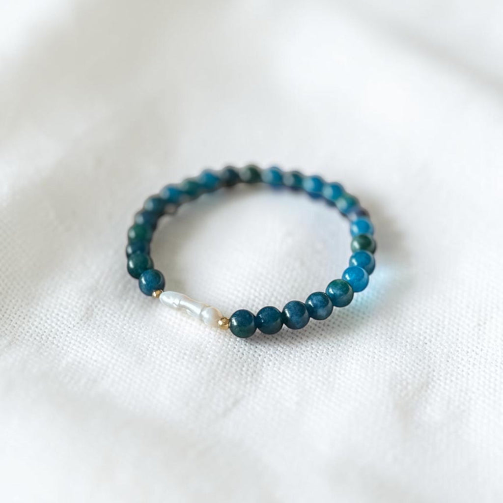 A beaded stretch bracelet featuring dark teal-blue round beads, a small white pearl-like bead, and a tiny gold spacer, resting on a soft white textured fabric background.