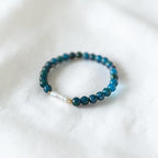 A beaded stretch bracelet featuring dark teal-blue round beads, a small white pearl-like bead, and a tiny gold spacer, resting on a soft white textured fabric background.