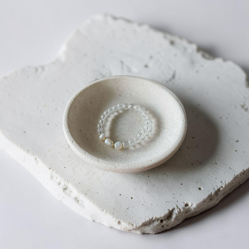 A delicate beaded bracelet with small white beads and clear crystal-like links, displayed inside a small round ceramic dish on a textured light gray stone slab.