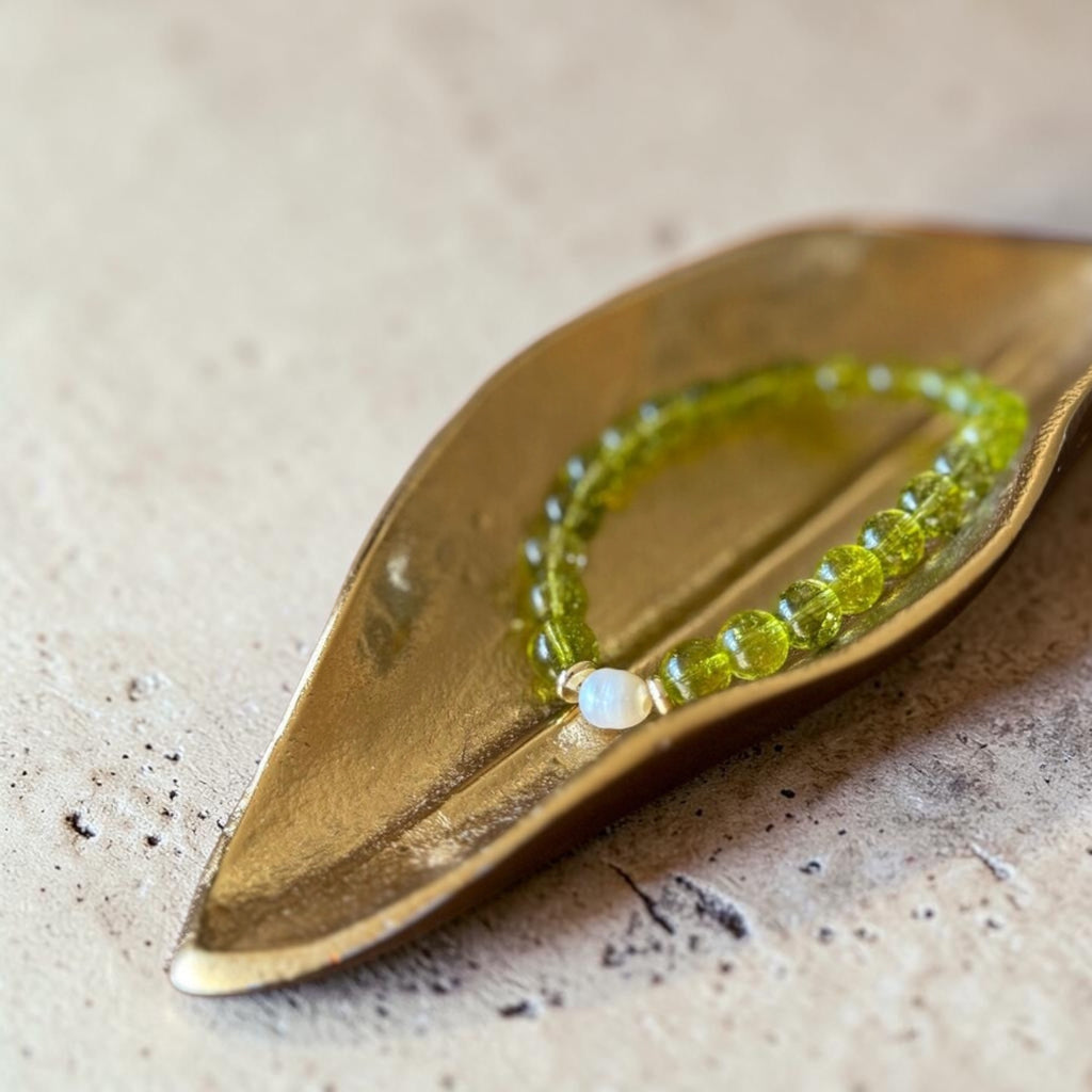 A beaded stretch bracelet in translucent olive green beads with a small white round stone accent, displayed on a polished gold leaf-shaped tray over a textured beige surface.