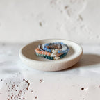 A stack of beaded bracelets in blue, peach, and white, displayed on a small round ceramic dish against a textured off-white background.