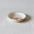 A delicate peach beaded bracelet with a silver-toned clasp resting inside a small, shallow white ceramic dish on a textured light gray surface.