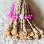A close-up of a beaded bracelet draped over a bundle of natural jute rope. The bracelet features bright pink translucent beads, a central white pearl-like bead, and small gold spacer beads, set against a neutral linen background.