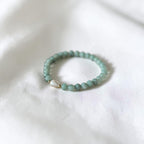 A beaded stretch bracelet made of light green beads with a small silver heart charm, resting on a white fabric background.