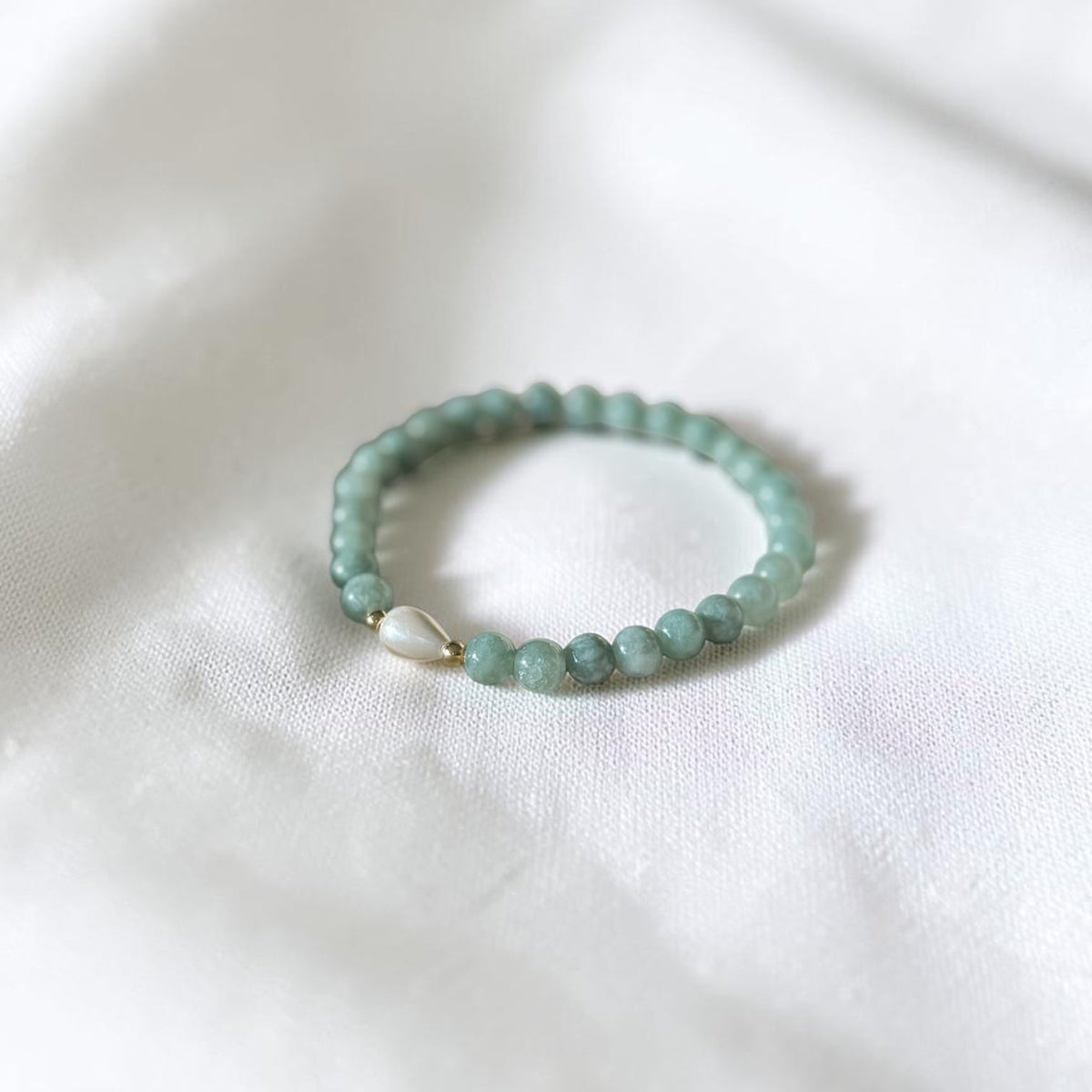 A beaded stretch bracelet made of light green beads with a small silver heart charm, resting on a white fabric background.